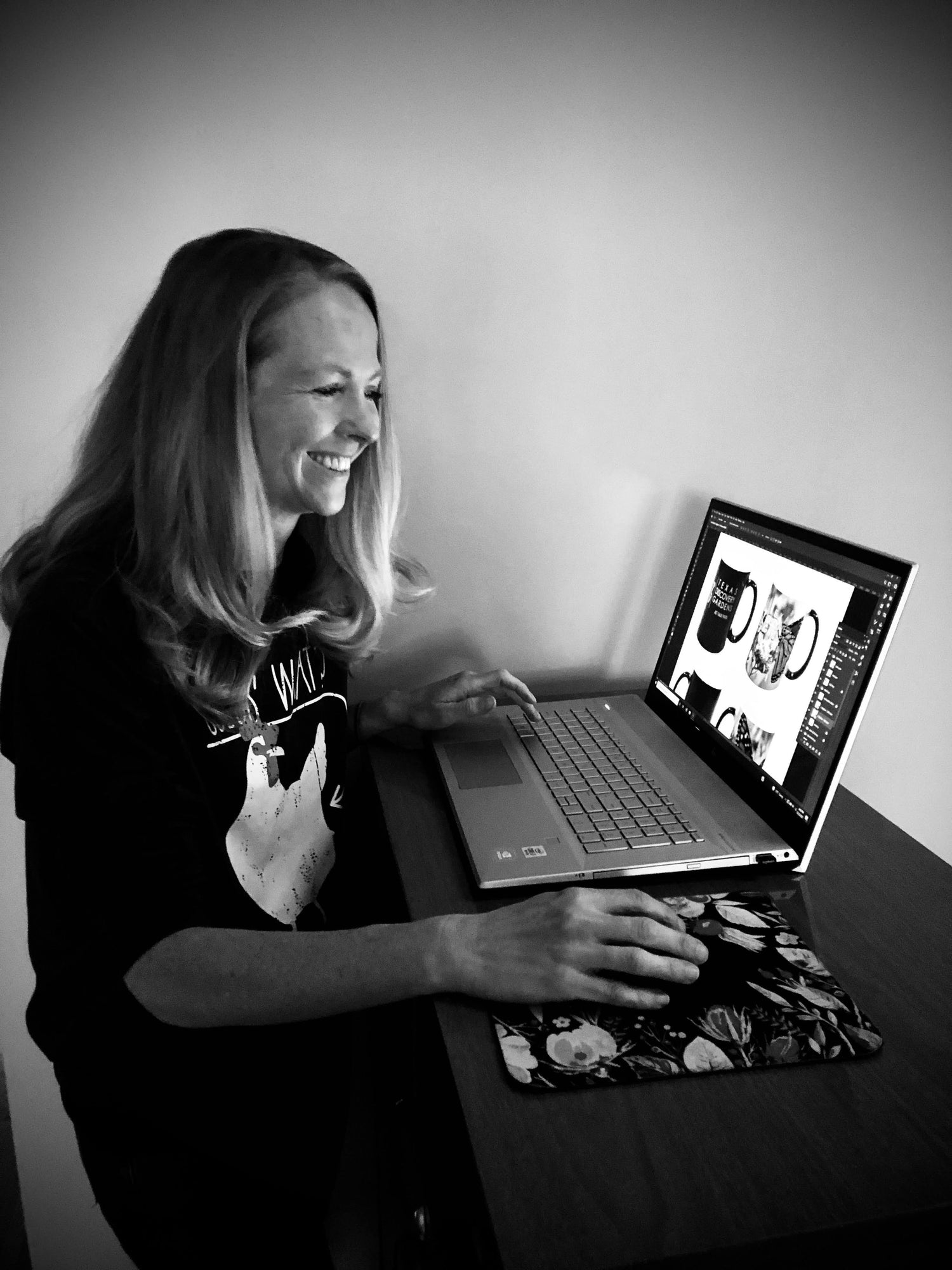 Smiling woman at desk with laptop working on designing mugs. 
